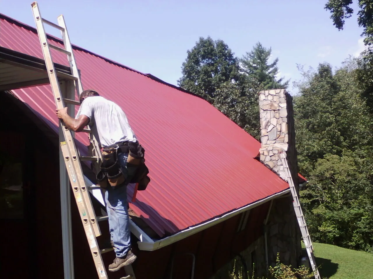 Roofer on a ladder beside a red metal roof and stone chimney — Soffit & Fascia Repair by Roofing Brothers in Seanor